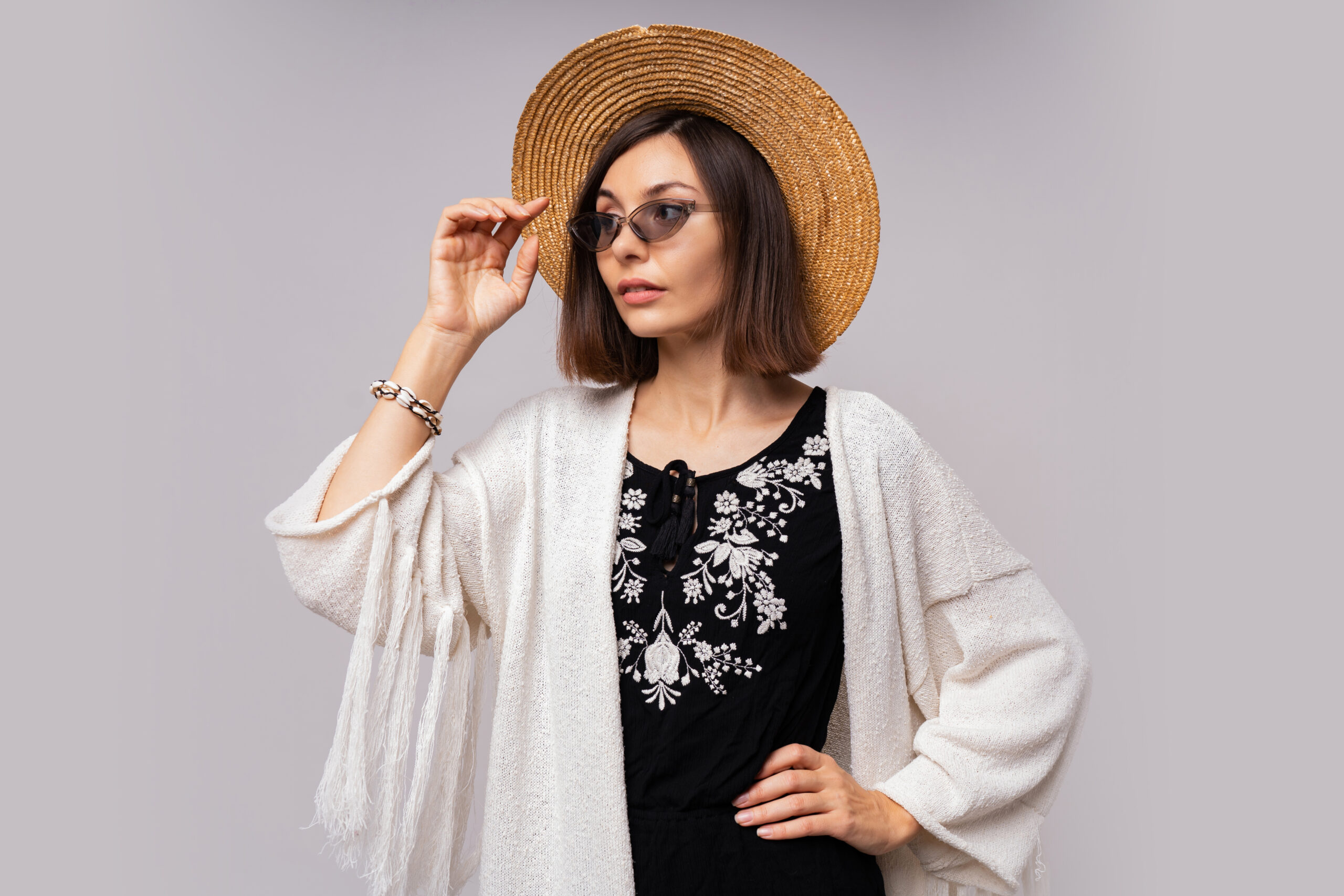 Debonair dark-eyed girl in straw hat and boho summer outfit posing on white background.